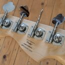 Detail hlavy basové kytary z přírodního světlého dřeva se čtyřmi chromovanými ladičkami. Na dřevě je černý nápis „Designed and Backed by Fender®“, po…