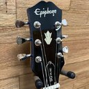 Černá hlava elektrické kytary s bílým nápisem Epiphone v horní části a bílým logem ve tvaru stylizovaného květu uprostřed. Na spodní části hlavy je s…
