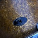 Detailní záběr na povrch bubnu s hnědo-zlatým kovovým efektem. Uprostřed je připevněn oválný černý štítek s nápisy REVELLE, WORLD'S SUPREME QUALITY a…