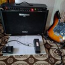 Černý kytarový kombozesilovač s nápisy BEHRINGER a V-TONE. Vedle něj stojí elektrická kytara s trupem v barevnosti sunburst, hnědým hmatníkem a tmavě…