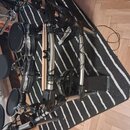 Elektronická bicí souprava s černými pady a kovovým chromovaným rámem, umístěná na černo-bílém pruhovaném koberci. Na jedné z horních trubek je vidit…