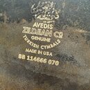 Detailní záběr na povrch mosazného činely s viditelnou patinou a oxidací. Na povrchu je vyražen nápis: AVEDIS ZILDJIAN CO GENUINE TURKISH CYMBALS MAD…