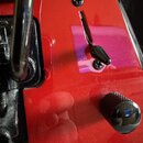 Detail červené elektrické kytary s kovovým mostem a černými ovládacími knoflíky. Na těle je vidět černý přepínač s kovovou pákou a dva připevněné šro…