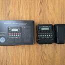 Černý kytarový procesor s digitálním displejem a řadou tlačítek. Na přístroji je uveden nápis G1X FOUR ZOOM. Vedle přístroje je umístěna jeho originá…