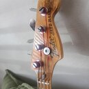 Fotografie zachycuje hlavu basové kytary z přírodního dřeva s lakovaným povrchem. Na hlavě je černý nápis „Fender® JAZZ BASS“ a pod ním menší, nečite…