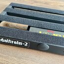Černý kovový pedalboard s nápisem pedaltrain-2 v bílém rámečku. Horní lišty jsou potaženy šedým protiskluzovým materiálem. Pod deskou je vidět část č…
