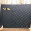 Černý kytarový kombozesilovač s pravou stranou pokrytou černou tkaninou s bílým kosočtvercovým vzorem. V horní levé části je umístěno zlaté logo VOX…