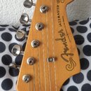 Fotografie zachycuje hlavu dřevěné elektrokytary v přirozené barvě. Na hlavě je černý nápis Fender, modelové označení JAZZMASTER a texty OFFSET Conto…