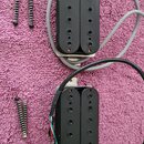Dvě černé humbucker snímače s kovovými základnami a připojenými kabely v šedé a černé barvě. Vedle snímačů jsou položeny čtyři montážní pružiny s šro…