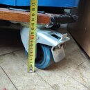 Fotografie detailně zachycuje modrý transportní case s dřevěnou hranou a kovovou pojízdnou kolečkem. Kolečko je z modrého plastu s kovovým držákem a…