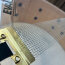 Detailní záběr na kovový snare wire (pružinu) v zlatavé barvě. Na kovovém držáku je vyražen nápis TAMA. Pružina je tvořena jemnou kovovou síťkou, kte…