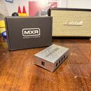 Na fotografii je stříbrný kovový napájecí adaptér s nápisem MXR dc brick a označením 18V DC IN. Vedle něj stojí černá kartonová krabice s bílým logem…