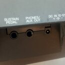 Detailní pohled na zadní panel šedého elektronického nástroje s třemi konektory. Nad vstupy jsou bílé nápisy „SUSTAIN PEDAL“, „PHONES/ AUX OUT“ a „DC…