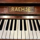 Detail klaviatury a horní části dřevěného pianina v hnědé barvě. Nad klávesami je umístěn nápis RAEHSE v zlatých uppercase písmenech na dřevěném podk…