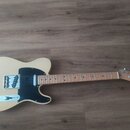 Elektrická kytara s krémovým tělem a černým pickguardem. Na hlavě hmatníku je vidět nápis Fender TELECASTER. Hmatník a krk jsou vyrobeny ze světlého…
