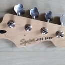 Fotografie zachycuje hlavu basové kytary z přírodního světlého dřeva se čtyřmi kovovými ladičkami. Na dřevě je černý nápis Squier JAZZ BASS® a pod ní…