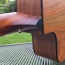 Detailní pohled na spoj hned krku a korpusu akustické kytary z dřeva s viditnou masivní strukturou. Nástroj má tmavé lemování po okrajích korpusu a k…