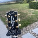 Fotografie zachycuje hlavu černé elektrické kytary s bílým lemováním a zlatými ladičkami. Na horní části je bílé logo Gibson a níže nápis Les Paul CU…