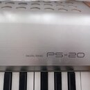 Detail digitálního piana s kovovým stříbrným povrchem a dřevěnými bočními listy. Na přední straně je černý nápis DIGITAL PIANO a vedle něj větší ozna…