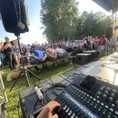 Fotografie zachycuje pohled z pódia na dav lidí před ní. V popředí je černý mixovací pult s řadou posuvníků a knoflíků, na kterém leží černý mobilní…
