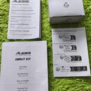 Fotografie zobrazuje bílé papírové návody a malou bílou kartonovou krabici na zeleném texturovaném povrchu. Na horním listu je logo ALESIS a nadpisy…