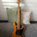 Elektrická basová kytara s přírodním dřevěným povrchem těla a hmatníkem. Na hlavě je vidět nápis Fender Jazz Bass. Instrument má černý pickguard a čt…