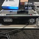 Na fotografii je kytarový pedalboard s několika zařízeními. V popředí je černý efekt s bílým logem MXR a bílým spirálovitým symbolem, nad ním je umís…