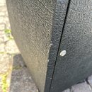 Detailní záběr na roh černého přístroje s texturovaným povrchem. Na hraně je vidět drobné odřasení a škrábance. V pravé části jsou vidět dvě stříbrné…
