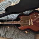 Elektrická kytara s tmavě červeným lakem a černým pickguardem v otevřeném tvrdém kufru. Na hlavě hmatníku je viditelné logo Gibson. Nástroj má dva hu…