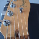 Záběr na zadní stranu hlavy kytary z přírodního dřeva. Na povrchu je vidět logo "Fender" a pod ním menší nápis "MADE IN USA". Jsou vidět čtyři kovové…