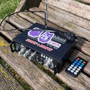 Černý audio zesilovač s kovovým šasi a dvěma bočními úchyty, umístěný na dřevěném stole. Na horní části jsou nalepené bílé štítky s nápisy „300W+300W…