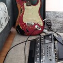 Elektrická kytara typu Stratocaster v červené barvě s výrazně odřeným povrchem, pod kterým je vidět dřevo. Na hlavě nástroje je nápis Fender a na bíl…