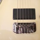 Detail elektrické kytary v krémové barvě. Je vidět černý humbucker a chromovaný kobylník s nápisem Ibanez. Povrch nástroje vykazuje v horní levé část…