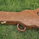 Hnědý tvrdý kufýr na kytaru s kovými zapínacími zámky a úchytem. Na pravé straně je vidět černé logo s nápisy USA a Gibson. Povrch vykazuje známky op…