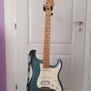 Elektrická kytara s modrým kovovým povrchem těla a světle hnědým javorovým hmatníkem. Na hlavě je viditelné logo Fender. Kytara má bílý pickguard, tř…