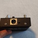 Fotografie zachycuje zadní stranu černého elektronického zařízení. Uprostřed je umístěn kovový konektor XLR s čtyřmi viditelnými šrouby. V horní část…