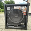 Černý basový kombozesilovač s kovovou ochrannou mřížkou přes reproduktor. Na přední straně jsou viditelná loga a nápisy: "Roland", "CUBE-120XL BASS",…