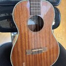 Hnědý akustický nástroj typu ukulele s lakovaným povrchem a bílým lemováním. Uvnitř ozvučnice je vidět štítek s logem a nápisem KALA. Nástroj má dřev…