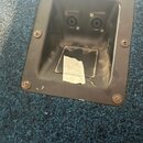 Fotografie zachycuje zapuštěný černý plastový panel s dvěma vstupy typu jack. Panel je přišroubován šesti šrouby do modrého texturovaného povrchu, pr…