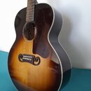 Akustická kytara s tmavě hnědým sunburst povrchem a bílým lemováním. Nástroj má tmavý hmatník, tmavě hnědý plechtr zaštitník a černý kobylník. Na těl…