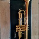 Zlatý mosazný trumpet položený na černém kufru. Na kufru je bílý nápis YAMAHA v obdélníkovém rámečku. Nástroj vykazuje známky používání v podobě drob…