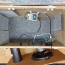 Fotografie zachycuje vnitřek otevřeného reproduktorového boxu s šedou akustickou výplní. Uvnitř jsou vidět elektroinstalace, transformátor a několik…