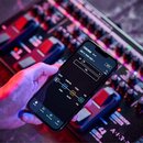 Ruka drží chytrý telefon s aplikací pro ovládání efektového pedálu. Na displeji je vidět logo BOSS, označení MC-90 a název presetu [001-1] ROCK LEAD.…