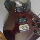 Elektrická kytara s tmavě červeným metalizovaným povrchem a dvěma černými humbuckery. Na kovovém mostku je vyryt nápis ALIEN WORKSHOP. Nástroj má tři…