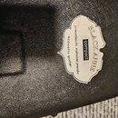 Detail černé texturované povrchové vrstvy pouzdra. Na povrchu je nalepená bílá štítková nálepka s nápisy BLACK ANNE, compact a acoustic guitar. Mezi…