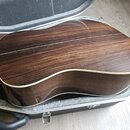 Zadní strana akustické kytary s tmavě hnědým dřevěným povrchem a výraznou sletovanou strukturou. Kolem hran korpusu je vidět světlý lem. Nástroj leží…
