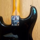 Zadní strana černé elektrické kytary s krkem z jasného dřeva. Na kovovém spojovacím plechu je vyryto logo Fender a nápis CORONA CALIFORNIA. V dolní č…