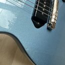 Detailní pohled na modrou elektrokitaru s kovovým leskem. Je vidět černý snímač typu humbucker a chromovaný most s saddles. Povrch laku vykazuje drob…