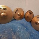 Čtyři mosazné činely značky Paiste v různých velikostech. Na činelech jsou čitelné nápisy „paiste 2002“ a konkrétní označení: „20" RIDE“, „16" CRASH“…
