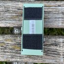 Fotografie zachycuje spodní stranu světle zeleného kovového efektpedálu. Na povrchu jsou přilepeny dva široké černé velcro pásy. Mezi nimi je umístěn…